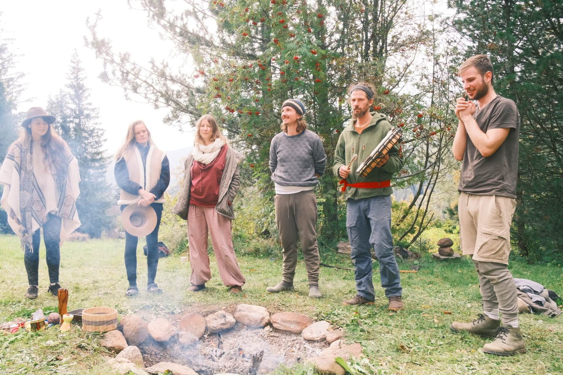 Sechs Menschen stehen im Kreis um eine rauchende Feuerstelle am Waldrand